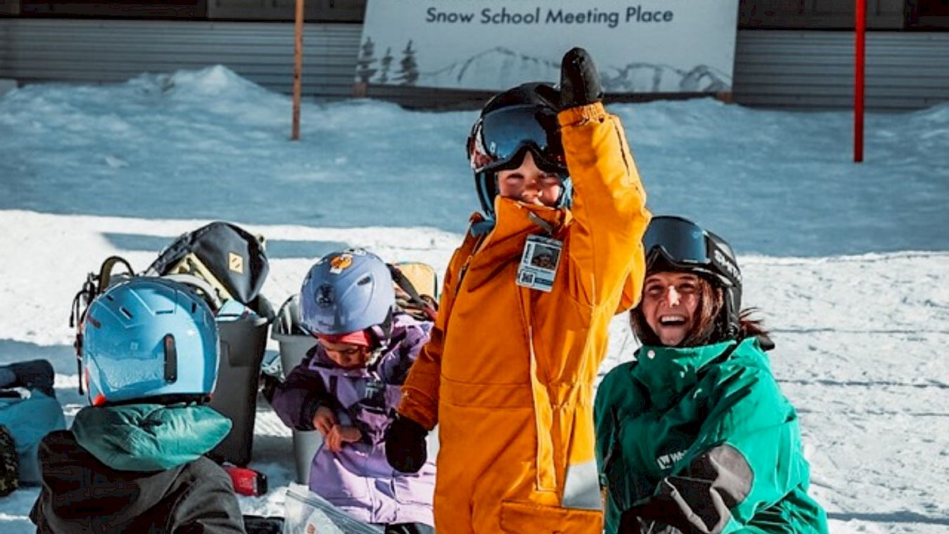 Girl in blue jacket learning to snowboard on the bunny hill at Whitewater while an instructor is nearby